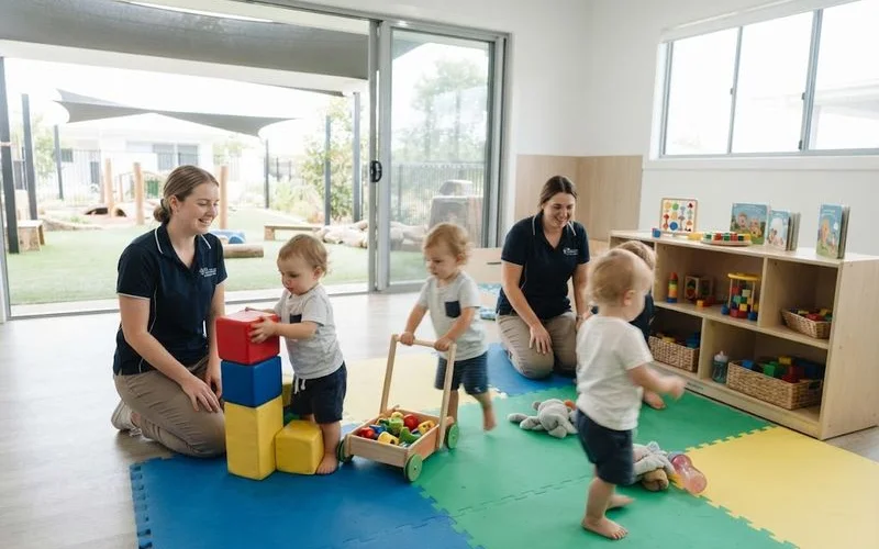 Two early childhood educators supervising a small group of toddlers during indoor floor play at a childcare centre